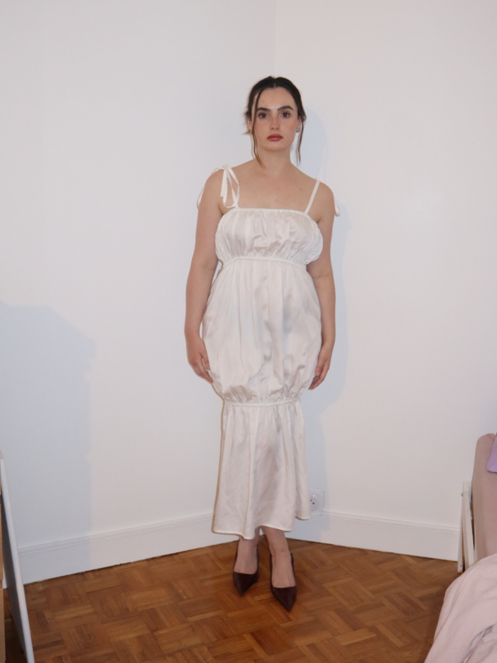 Woman wearing a white dress standing in a room with wooden flooring and a white wall.