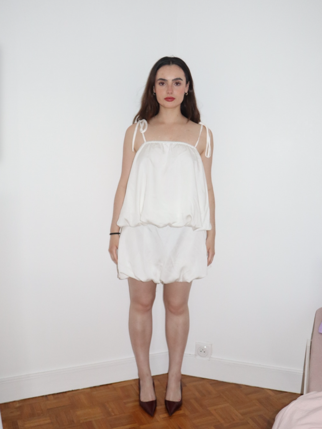 Woman wearing a white dress standing against a plain wall.