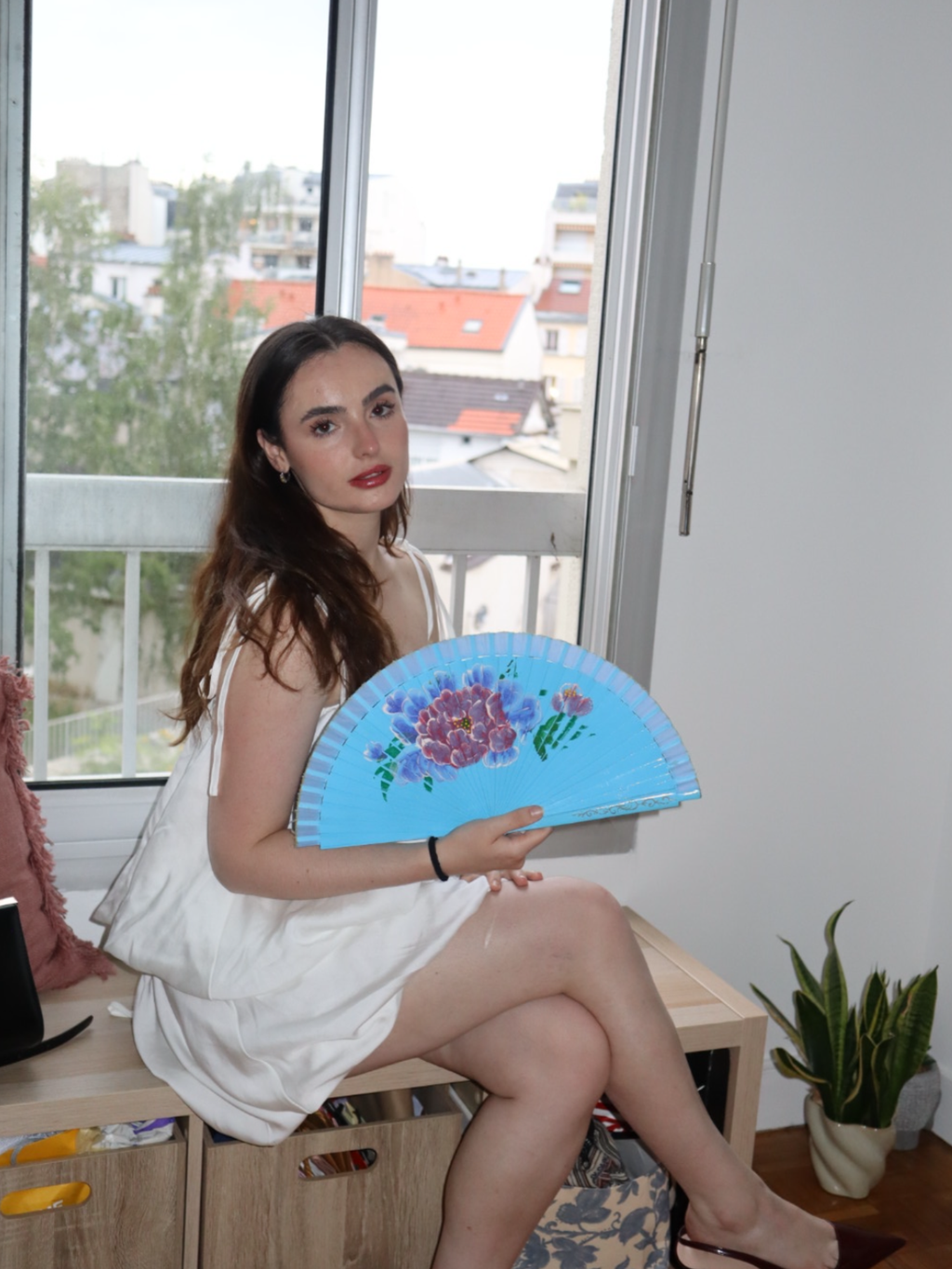 Woman holding a blue floral fan in a room with a window view
