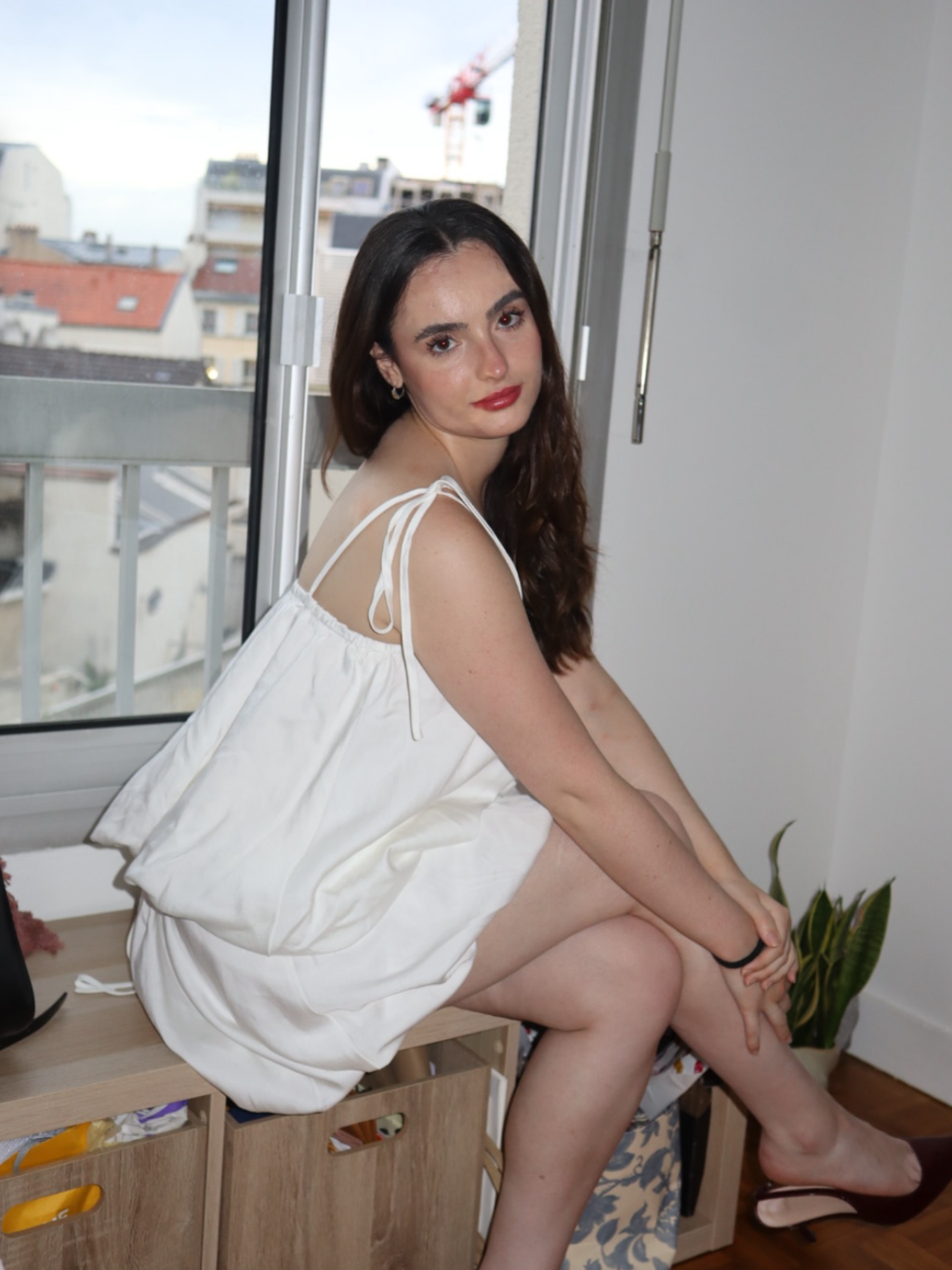 Woman in a white dress sitting on a window sill with a cityscape view.