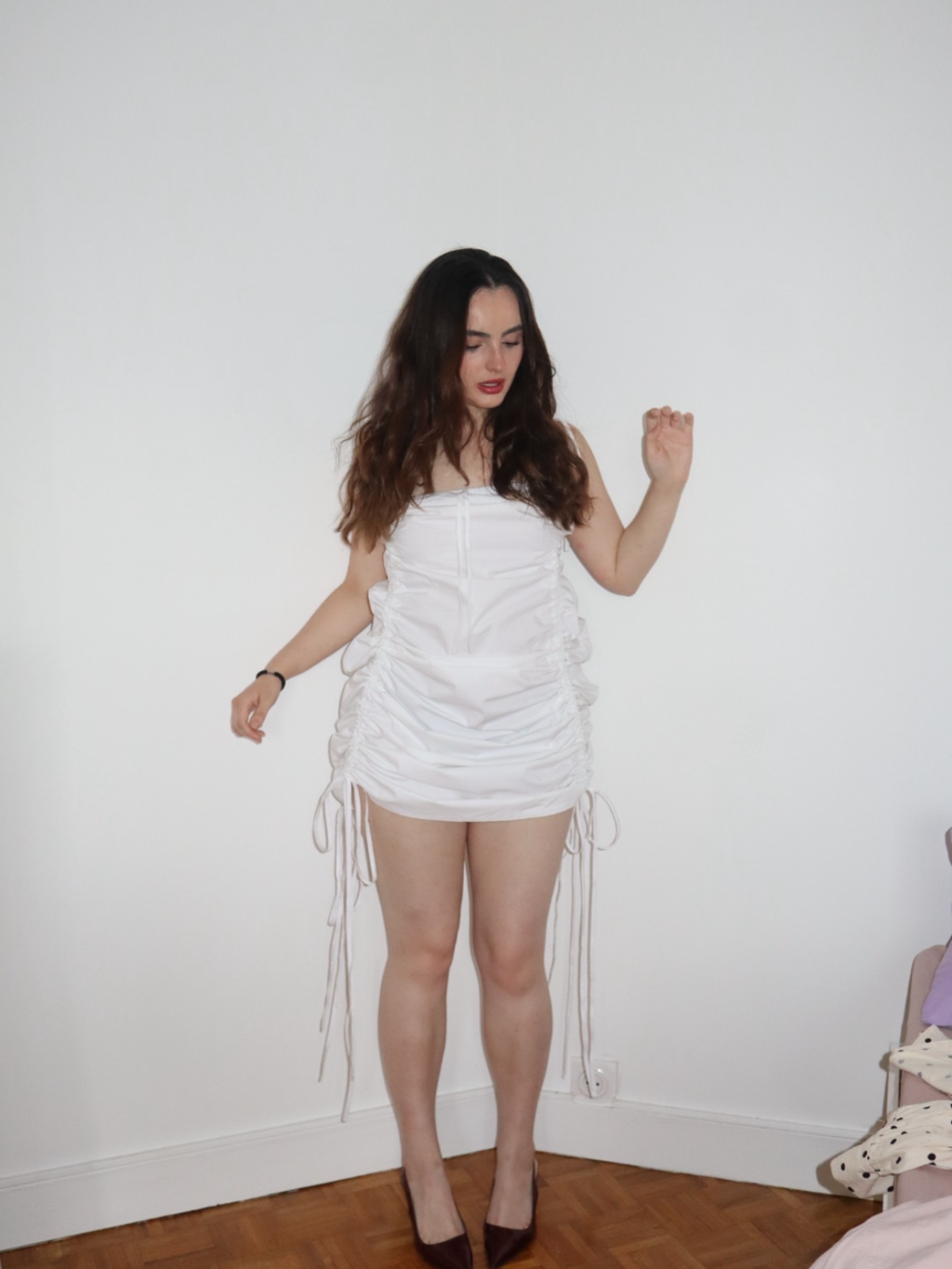 Woman wearing a white dress with ruched details against a plain white wall.